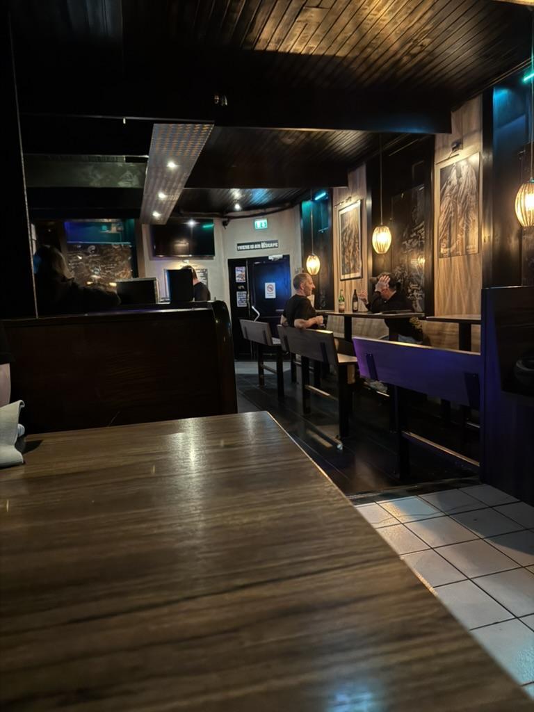 The image depicts the interior of a dimly lit bar or lounge. The floor is a mix of dark wood and white tiles, receding into the distance along a central aisle. Several people are seated at dark wood tables and a bar counter, with a doorway visible at the back of the room. The ceiling is a dark wood paneling, and there are hanging pendant lights illuminating the space. On the back wall, there is a sign that reads "THERE IS AN EXIT.”