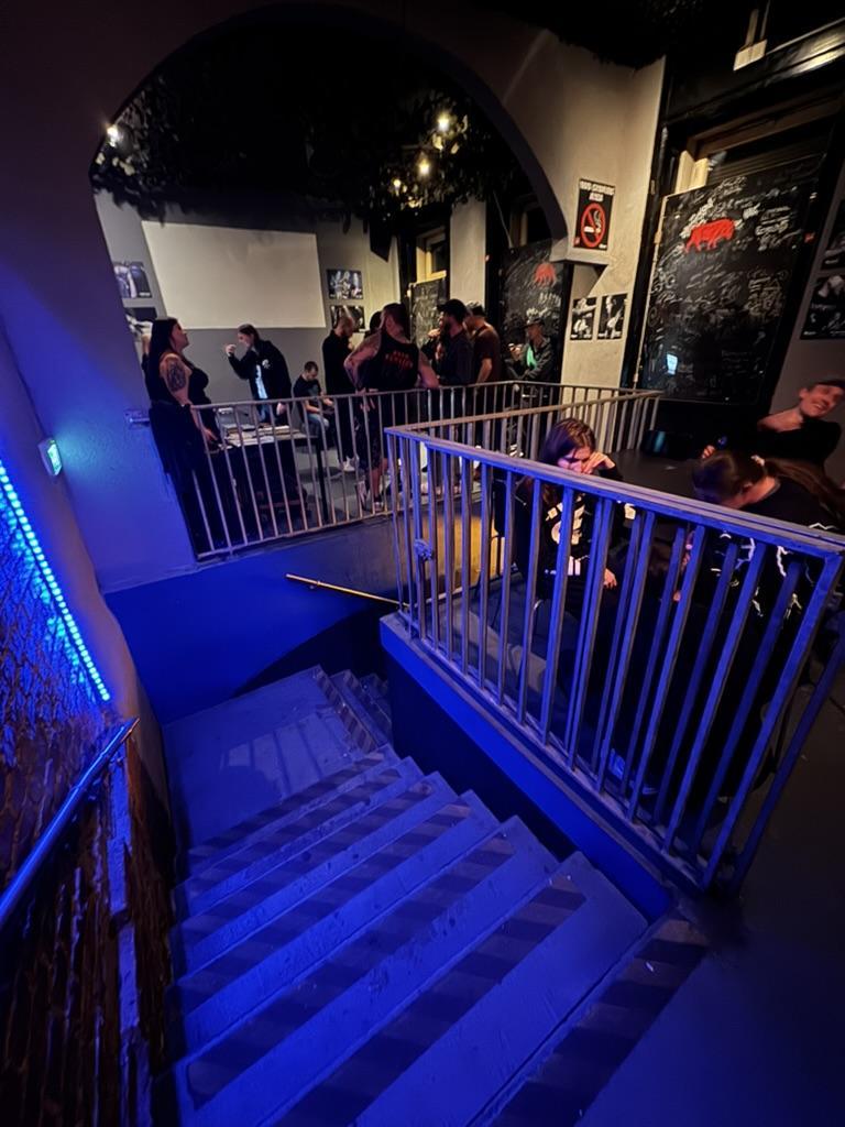 A dimly lit interior space is shown from a high angle, looking down a set of wooden stairs. The stairs are flanked by metal railings, illuminated with bright blue lighting. A crowd of people are gathered at the top of the stairs, partially obscuring the view of the space beyond, which appears to be a bar or performance area. Posters and artwork adorn the walls on either side of the stairs. A sign visible on the right wall reads "NO RE-ENTRY”