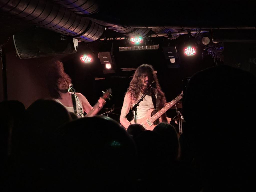 The image depicts a live musical performance taking place on a dimly lit stage. Two figures are visible in the foreground, both playing guitars. The person on the left is partially obscured and holding a guitar, while the person on the right is facing forward, singing into a microphone while also playing a guitar. A crowd of people is visible as dark silhouettes in the very foreground. The stage is lit by several colorful lights, primarily red and green, mounted on the wall behind the performers.