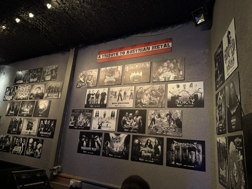 A dark gray wall is completely covered in framed black and white posters. The posters appear to be photos of various musical groups and individual performers, likely bands. They are arranged in a grid-like pattern, with multiple rows and columns filling the entire visible wall space. At the very top center of the wall, a rectangular sign reads, **"A TRIBUTE TO AUSTRIAN METAL"** in white lettering. Along the very bottom edge of the wall are dark, upholstered seats and a bit of a person's head.